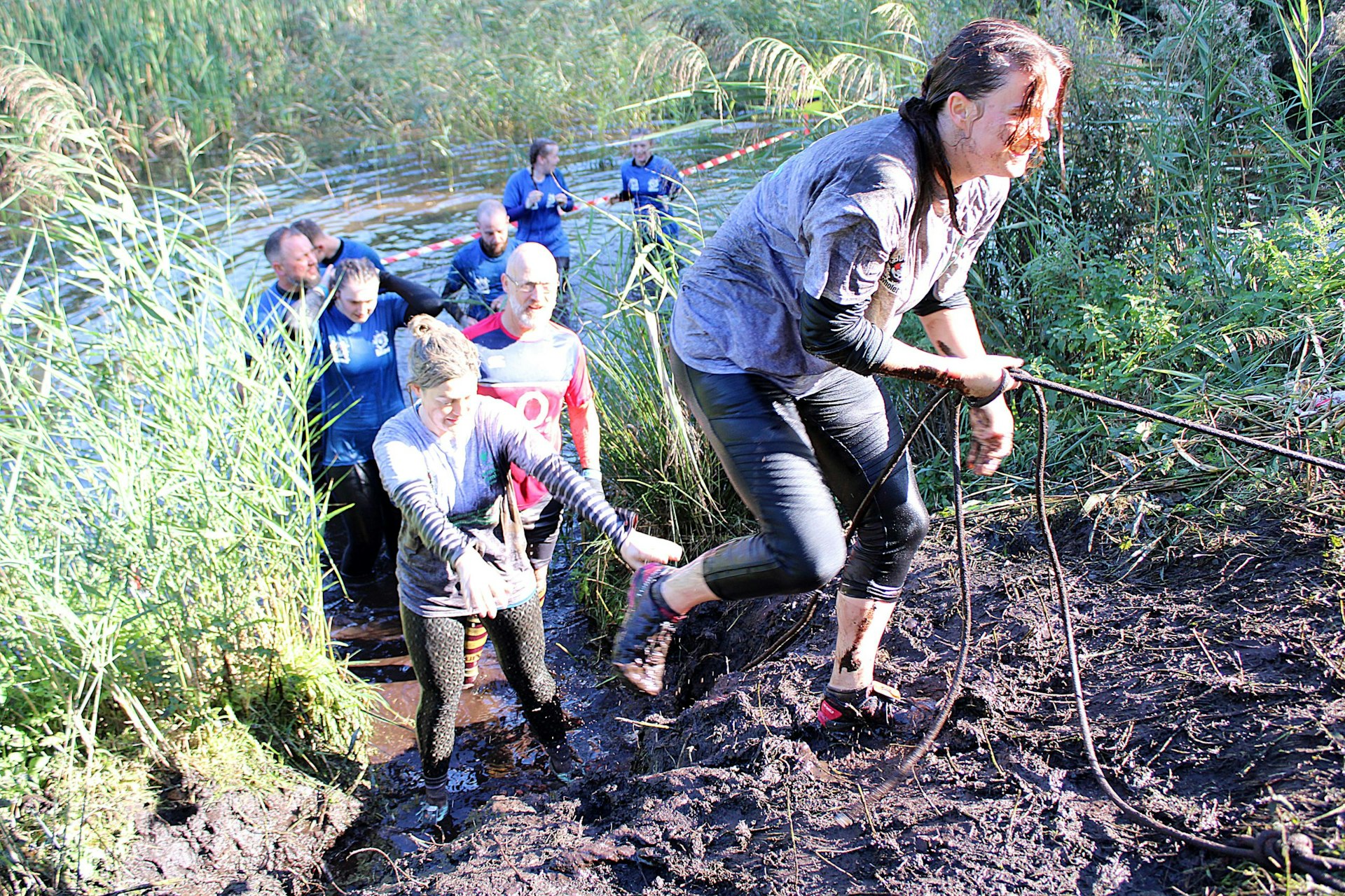 Group of Whole Hog volunteers climbing out of a muddy water pit using a rope during the obstacle race, smiling and supporting each other